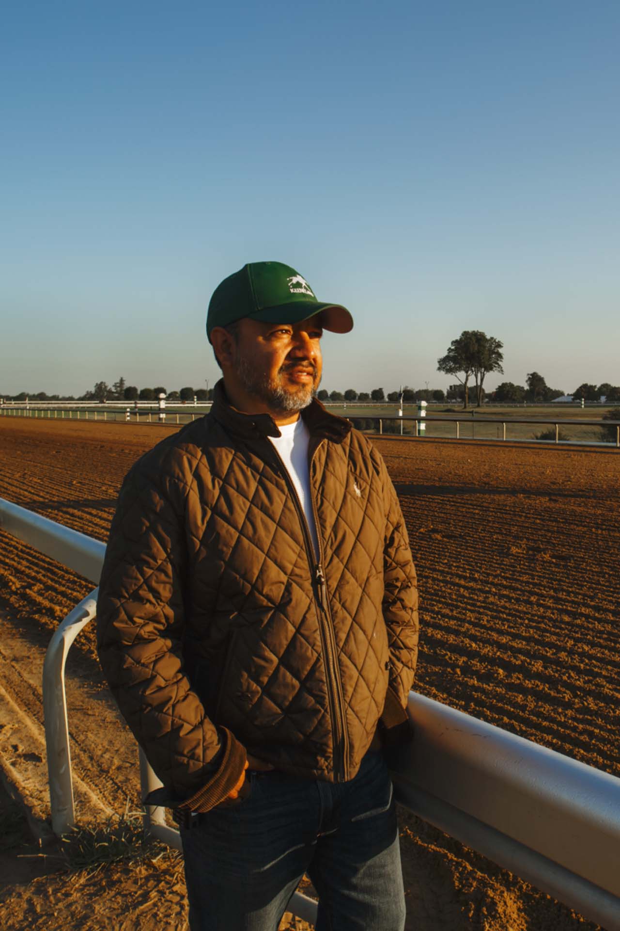 A photo of Alfredo Laureano looking off into the distance while standing by the dirt track at sunset. He is an older Hispanic man with graying chin stubble. He is wearing a green Fasthorsepro-brand hat and a brown Fasthorsepro-brand jacket overtop a white shirt.
